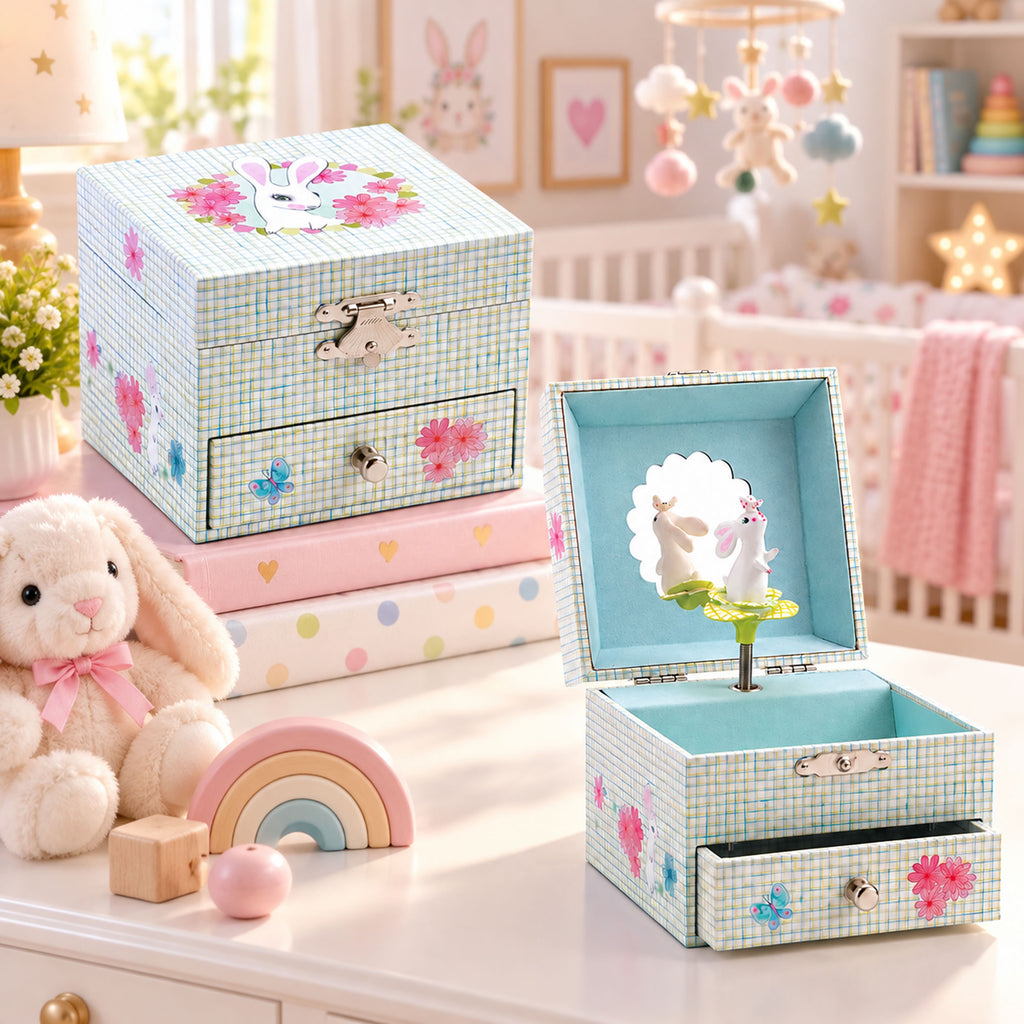 A decorative baby nursery jewelry box set displayed on a white dresser in a softly styled nursery. The set includes a closed light blue gingham box with pink flowers and a bunny motif, a pink polka-dot storage box, and an open matching box with a rabbit figurine inside and a small tree decoration.