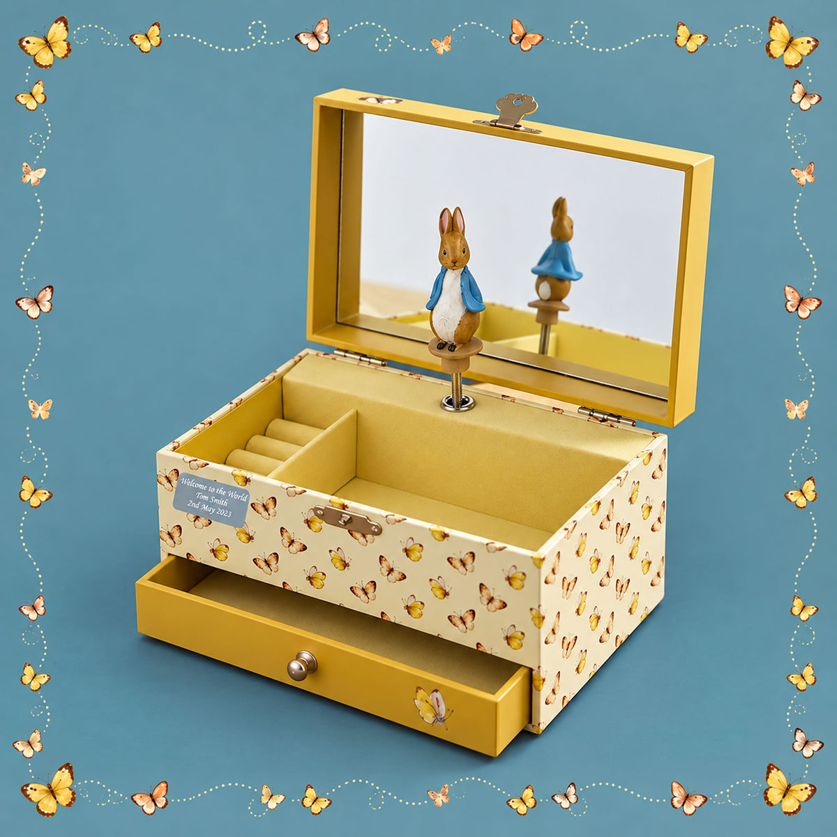 A Peter Rabbit-themed wooden jewelry box with a yellow interior and exterior patterned with small rabbit illustrations. The top lid features a mirror reflecting a rabbit figure, and the bottom drawer is pulled out.