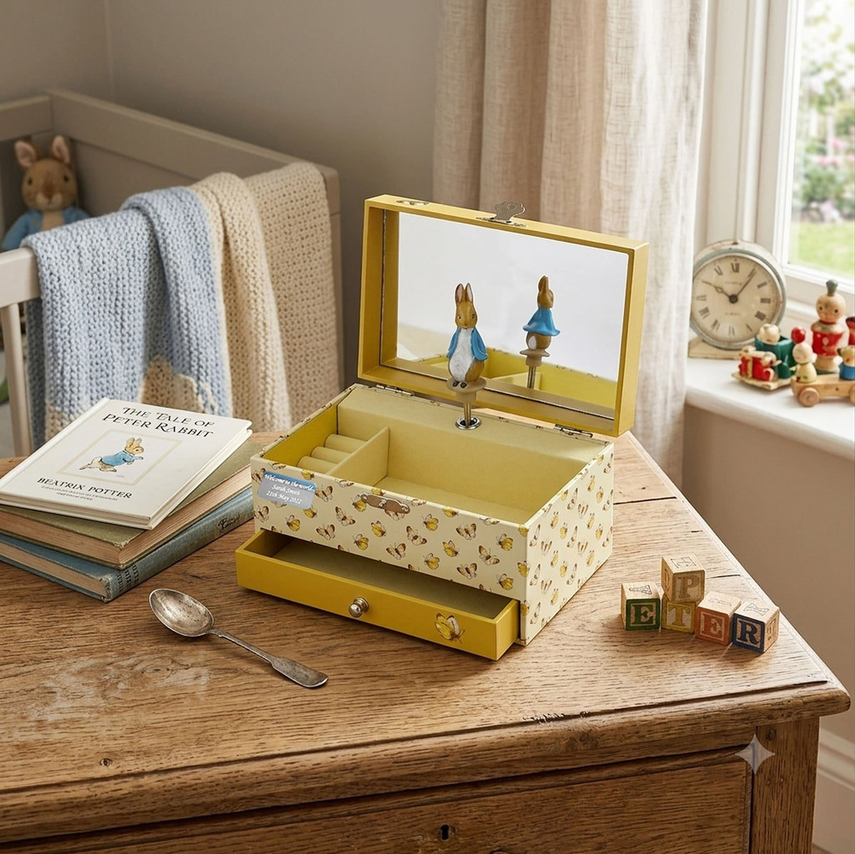 A yellow Peter Rabbit-themed music box with an open mirror lid, displayed on a wooden table in a child's bedroom setting next to a book and toy blocks.