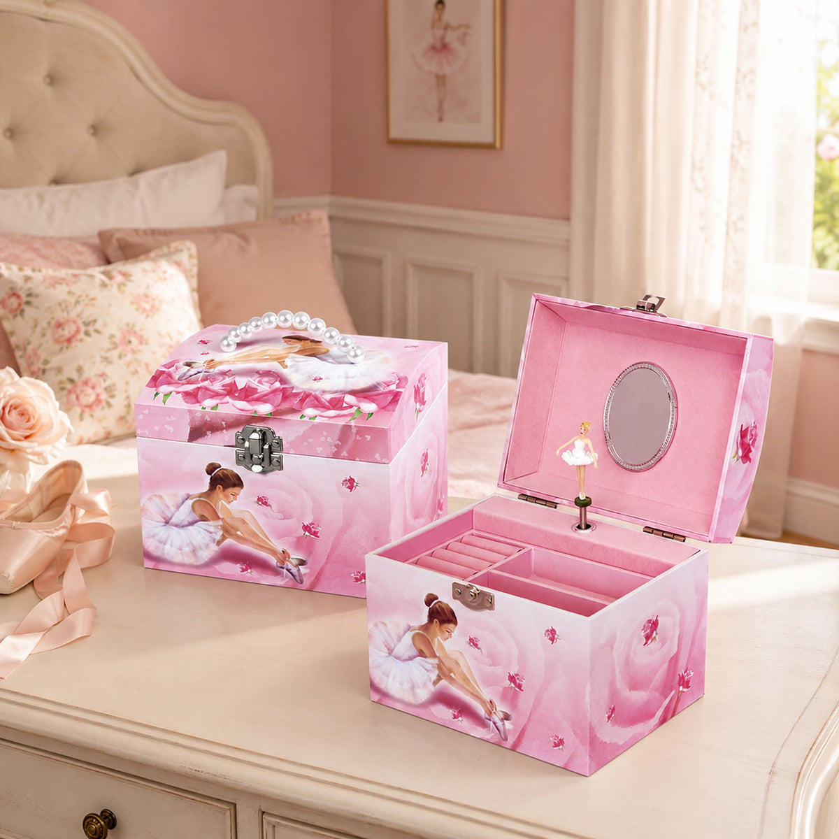 A pink ballerina-themed kids' jewellery box set is shown in a bedroom setting. One box is closed, featuring a silver clasp, a decorative handle, and a printed ballerina illustration.