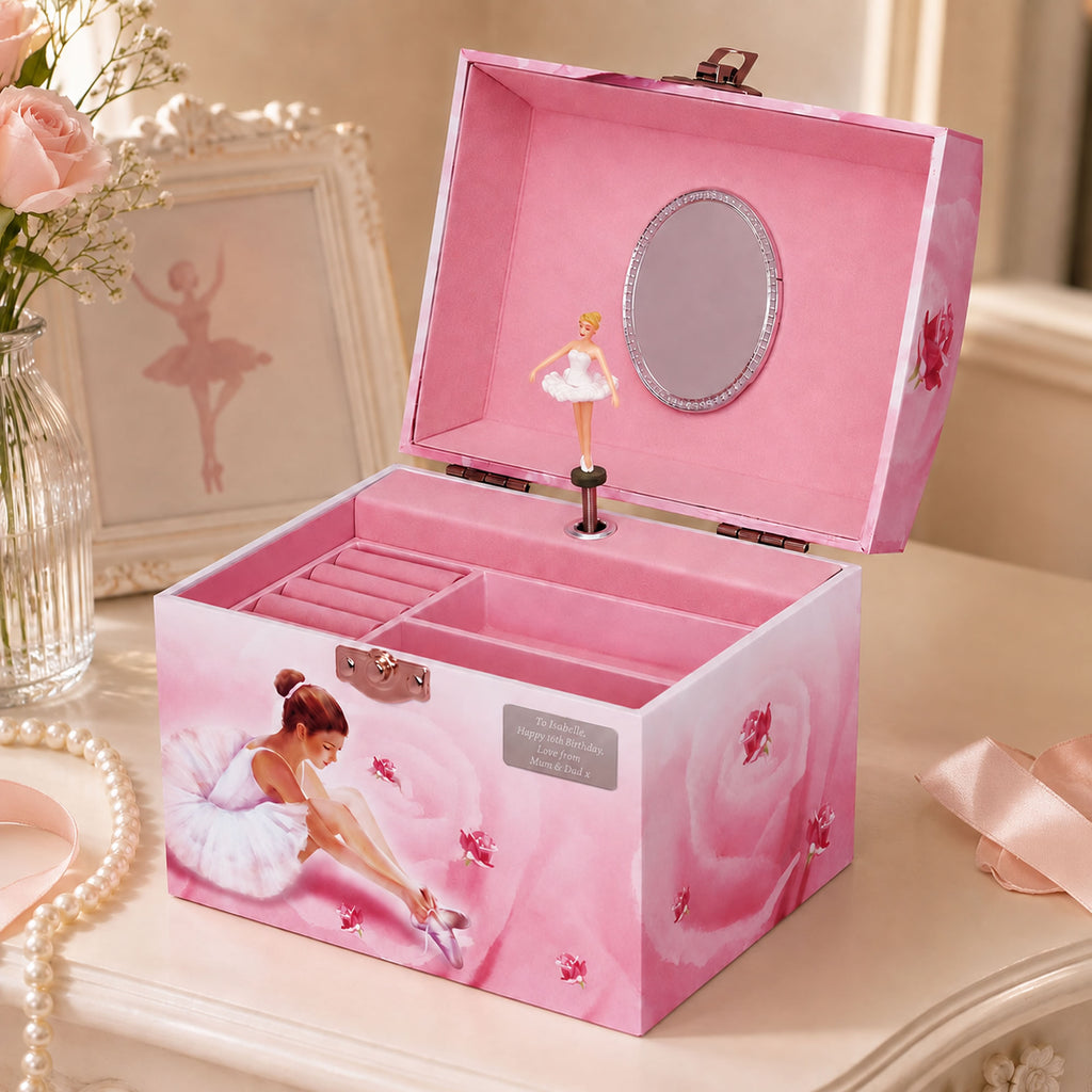 A personalised pink ballerina-themed jewellery box with a hinged lid, open to reveal a small ballerina figurine, mirror, and interior compartments. The exterior is decorated with pink roses and an illustration of a ballerina sitting on a chair, shown on a light-colored table with decorative props.