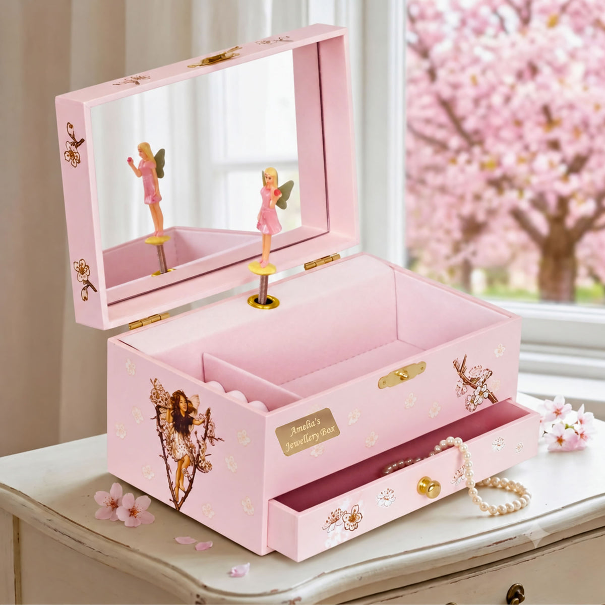 A pink fairy-themed jewelry box with an open lid showing a mirror and two small fairy figurines, placed on a white table with cherry blossoms in the background. A pearl necklace is draped over the front drawer.