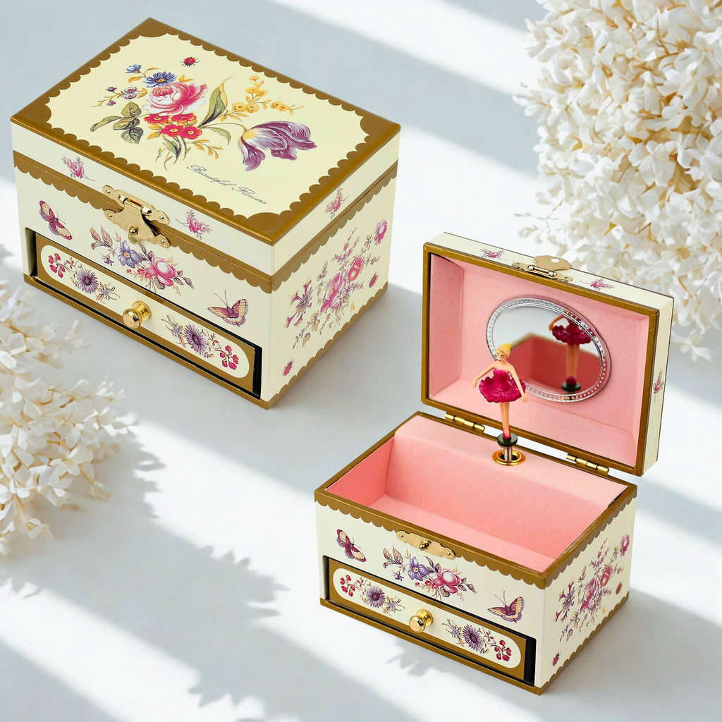 Two decorative music boxes with floral prints and gold trim, one closed and one open showing a pink interior and miniature ballerina, styled with white flowers on a white background.