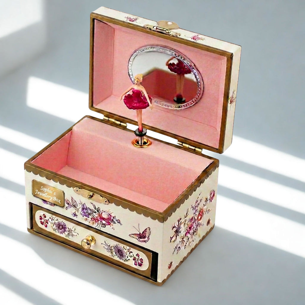 An open decorative music box with a pink interior, floral and butterfly illustrations on the exterior, a small mirror in the lid, and a ballerina figurine on a stand, photographed on a light surface with soft window shadows.