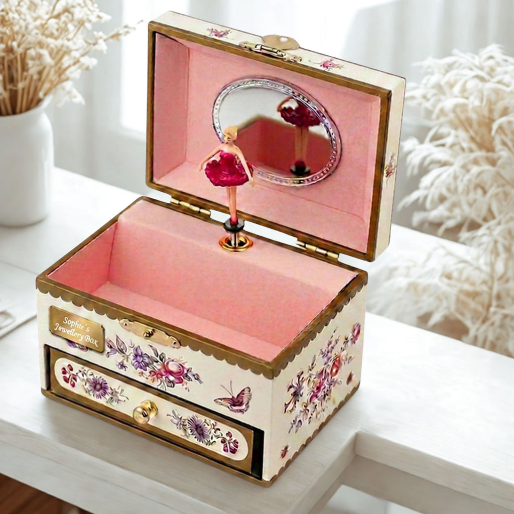 A decorative music box jewelry box with a pink interior, gold trim, floral and butterfly illustrations, a small ballerina figurine, and an open lid mirror, displayed on a white table.
