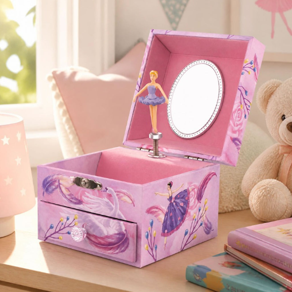 A pink music box jewelry box with ballerina illustrations, shown open on a wooden table with a teddy bear, books, and a lamp in a softly lit room.