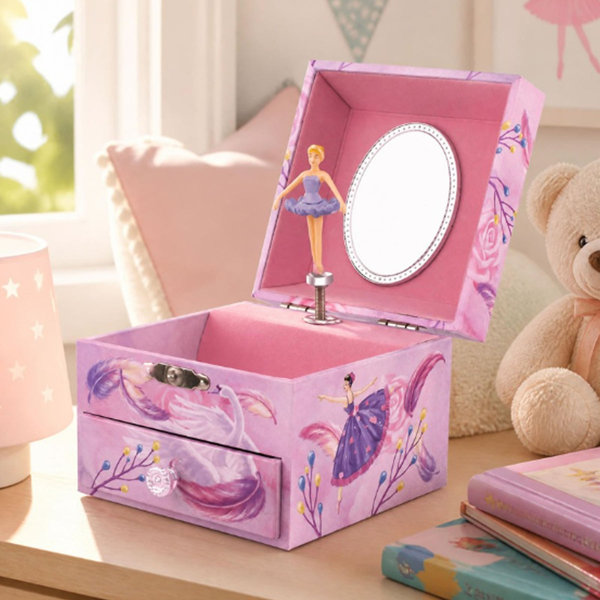 A pink music box jewelry box with ballerina illustrations, shown open on a wooden table with a teddy bear, books, and a lamp in a softly lit room.
