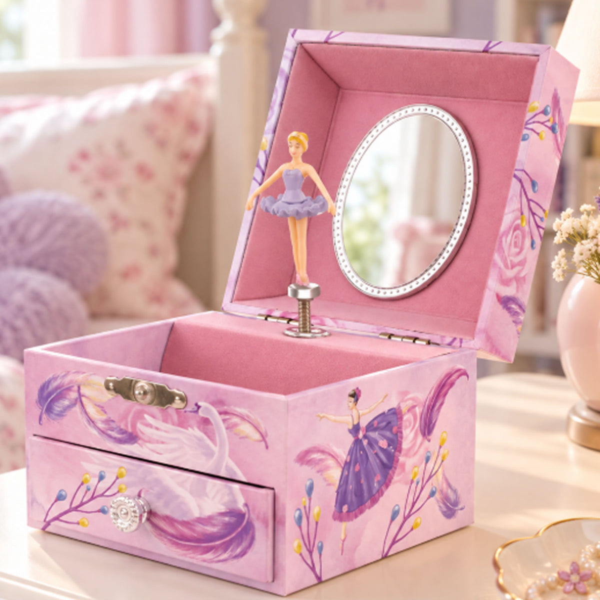 A pink ballerina-themed jewelry box with a hinged lid, open to reveal a small mirror and ballerina figurine. The box features painted ballerina and swan designs, a front drawer with a decorative clasp, and sits on a light-colored table in a softly blurred bedroom setting.