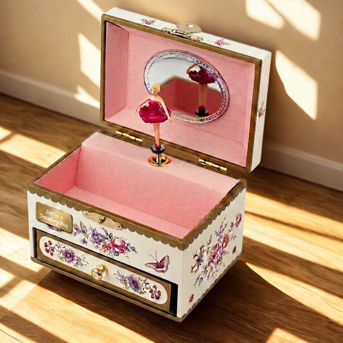 An open decorative music box with a pink interior, floral and butterfly illustrations on the exterior, a small mirror in the lid, and a ballerina figurine on a stand, placed on a wooden floor in warm sunlight.
