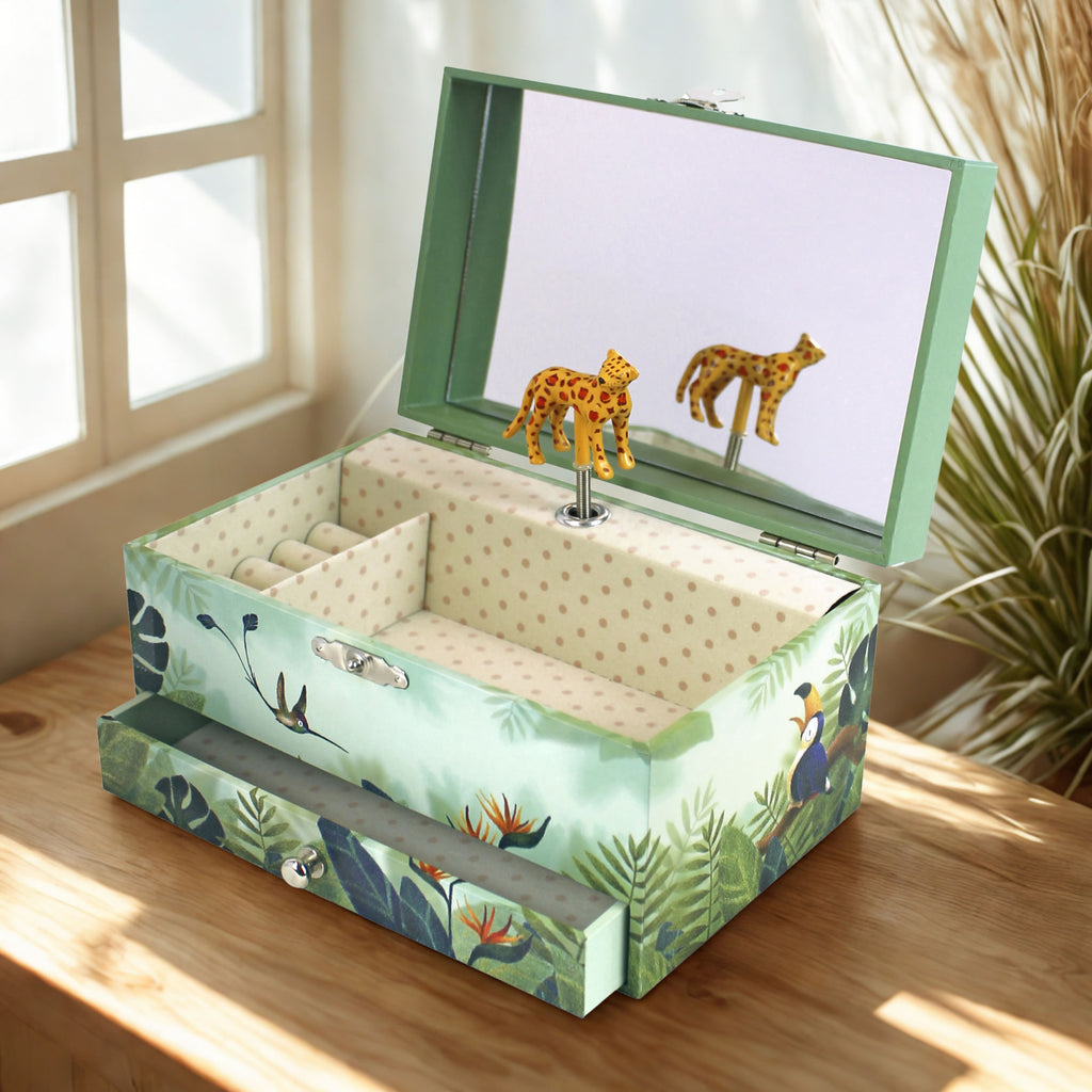 A decorative jewelry box with a tropical jungle print, shown open on a wooden table in a sunlit room. The box has a mint green frame, patterned exterior, and interior with gold polka dots.